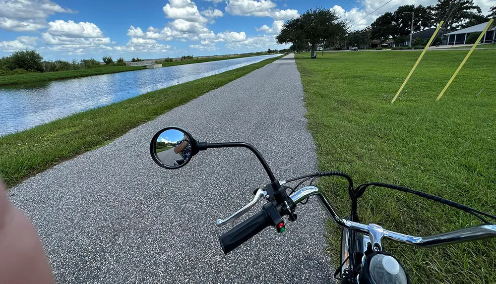 A persons view from a bicycle handlebar featuring a rearview mirror reflecting the cyclists face overlooking a paved path next to a waterway and greenery under a partly cloudy sky