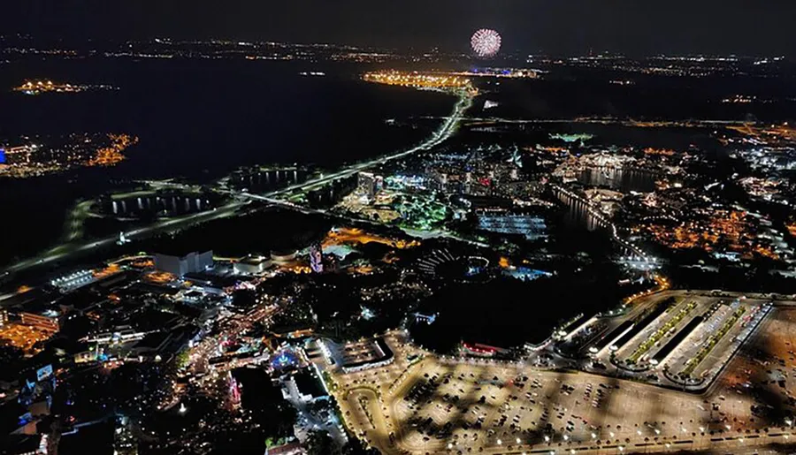 The image shows a night-time aerial view of a brightly lit urban area with a fireworks display in the background.