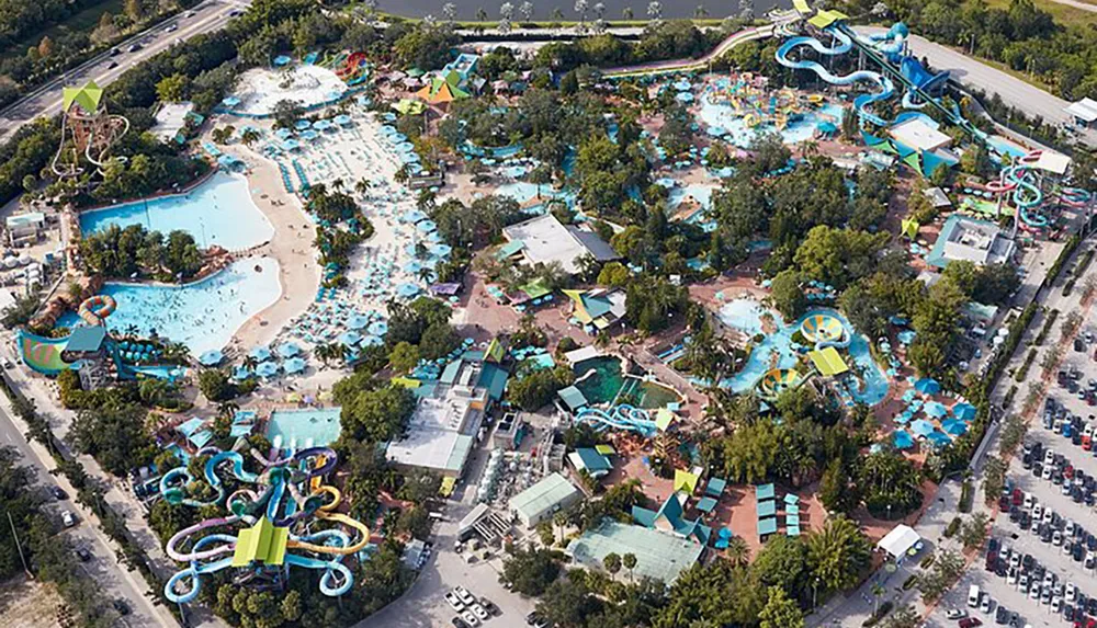 The image shows an aerial view of a colorful water park with a variety of slides pools and leisure areas surrounded by parking and greenery