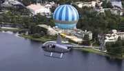 A helicopter is depicted flying close to a large, anchored hot air balloon over a scenic area with buildings and a body of water.