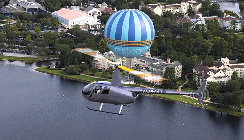 A helicopter is depicted flying close to a large anchored hot air balloon over a scenic area with buildings and a body of water