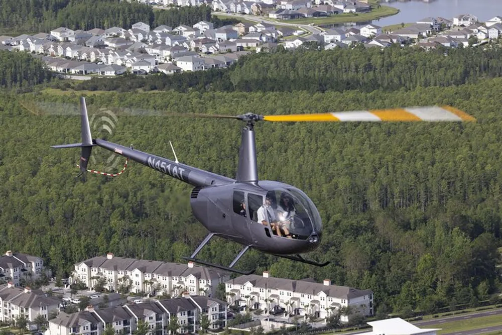 A helicopter is flying over a suburban area with a pilot and passenger visible in the cockpit