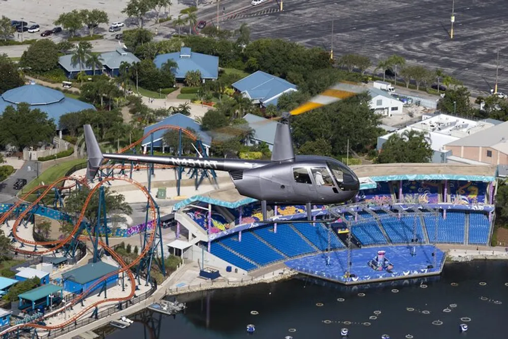 The image shows a helicopter flying over an amusement park with a roller coaster and stadium seating by a body of water