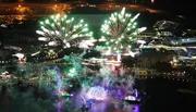 The image features a nighttime aerial view of a lively amusement park with vibrant fireworks bursting over lit-up attractions and bodies of water.