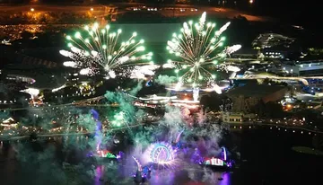 The image features a nighttime aerial view of a lively amusement park with vibrant fireworks bursting over lit-up attractions and bodies of water.