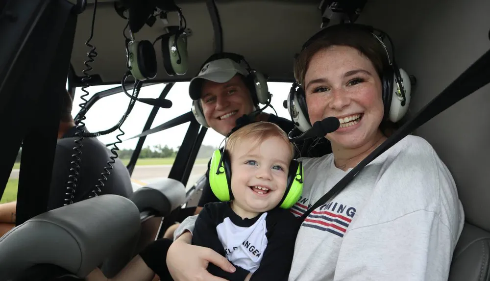 A family of three is smiling inside a helicopter each wearing headsets suggesting they are about to experience a flight together