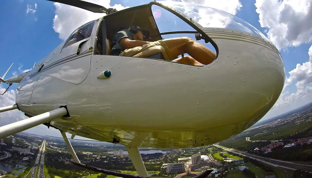 The image shows a close-up view of a helicopter in mid-flight with a pilot inside captured from an angle that presents the nose of the aircraft and the landscape below the clear sky