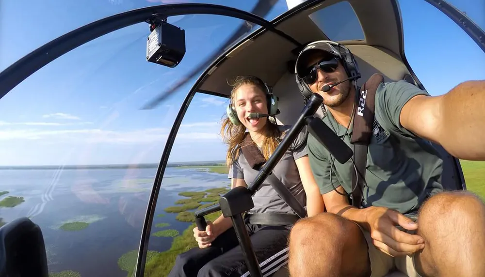 A man and a woman are smiling and taking a selfie inside a helicopter cockpit high above a landscape of water and greenery