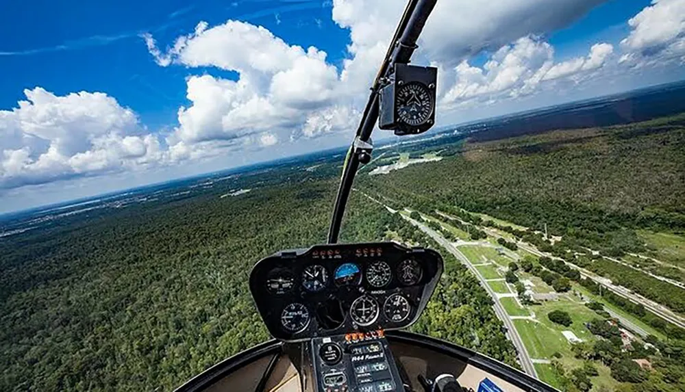 The image shows the cockpit view from a helicopter with its instrumentation visible and a landscape with roads and trees below