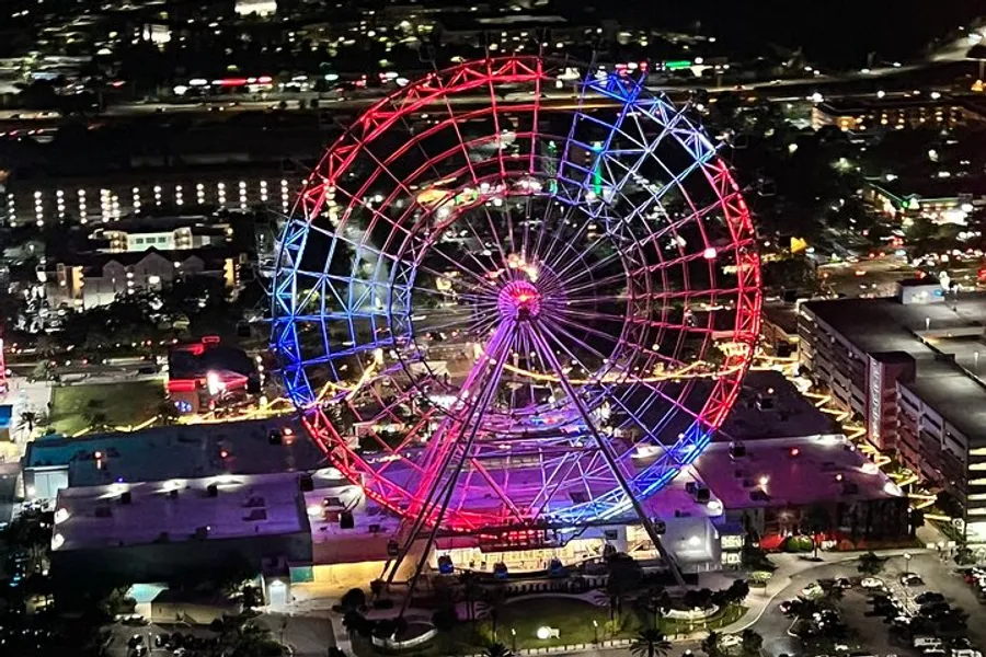 An illuminated Ferris wheel stands out against the night skyline, casting vibrant red and blue hues over the surrounding area.