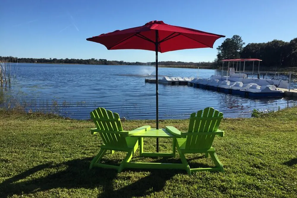 Two green Adirondack chairs and a matching table sit under a red umbrella facing a tranquil lake with a dock and pedal boats