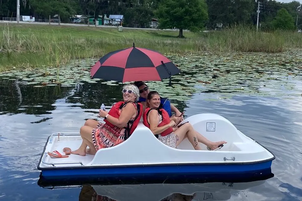 Three people are enjoying a sunny day on a pedal boat in a calm body of water with one person holding a red and black umbrella