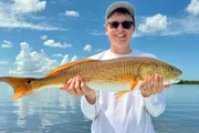 A person is smiling while holding a large fish in front of a body of water under a clear blue sky.