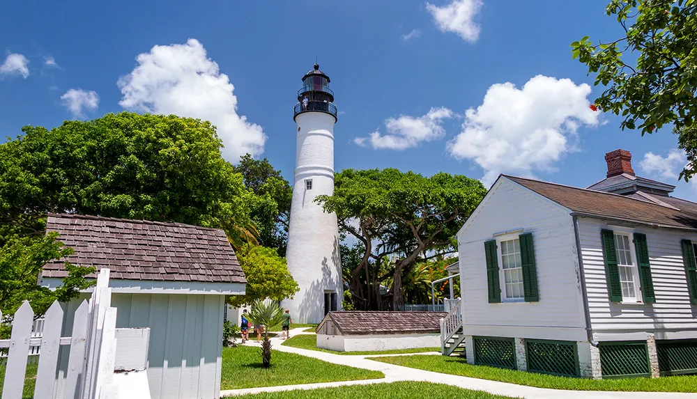 A classic white lighthouse stands tall amidst greenery under a blue sky flanked by traditional white structures with green shutters