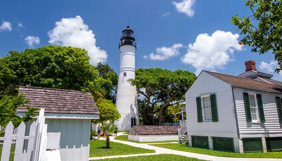 A classic white lighthouse stands tall amidst greenery under a blue sky, flanked by traditional white structures with green shutters.
