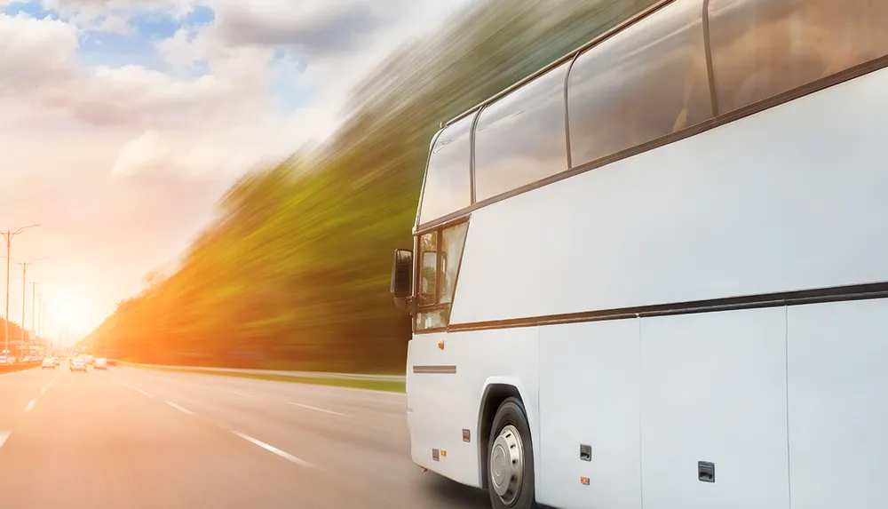 A white coach bus is shown in motion on a road with blurred scenery indicating high speed and the bright sun visible in the background suggesting either dawn or dusk