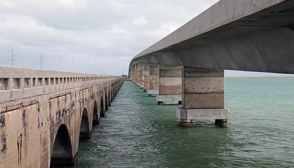 The image shows two parallel bridges over water extending toward the horizon with the older bridge on the left and the modern one on the right under a cloudy sky