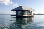 A small, cozy floating house is peacefully situated on a calm body of water under a clear sky.
