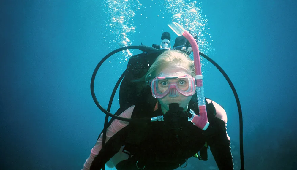 A scuba diver with a pink snorkel and mask is submerged in clear blue water surrounded by bubbles