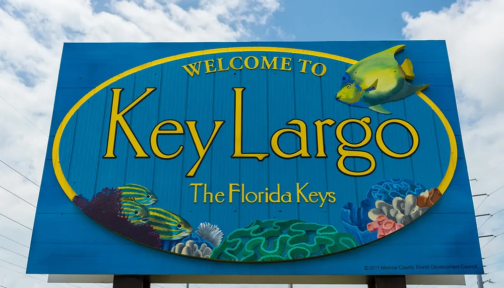 The image shows a colorful welcome sign with the text Welcome to Key Largo - The Florida Keys surrounded by marine life illustrations against a blue sky with clouds