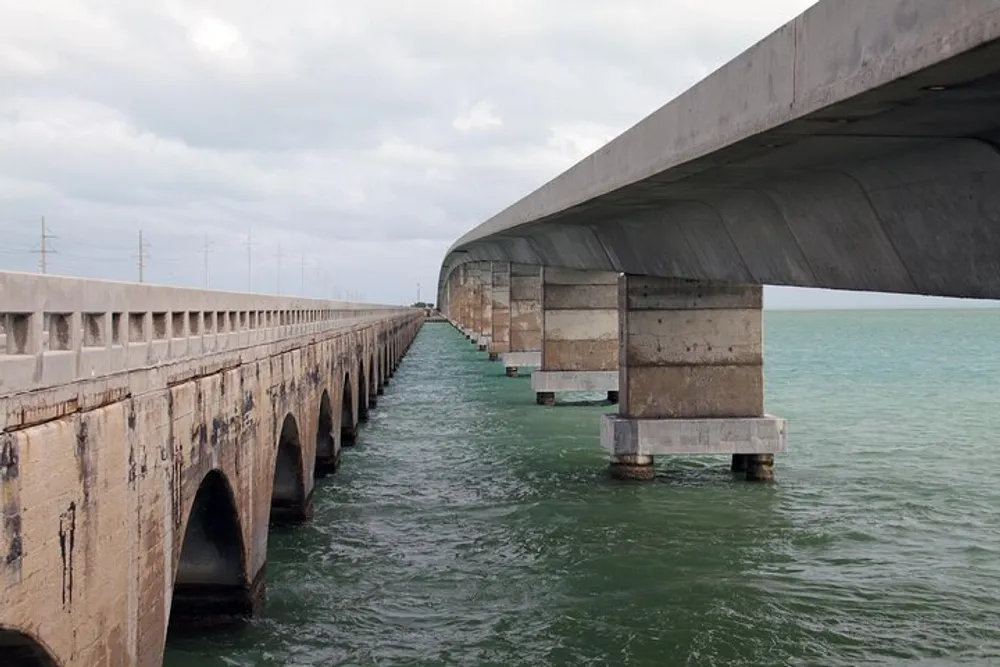 The image shows two parallel bridges extending over a body of water with the older bridge appearing weathered and the newer one looking more robust