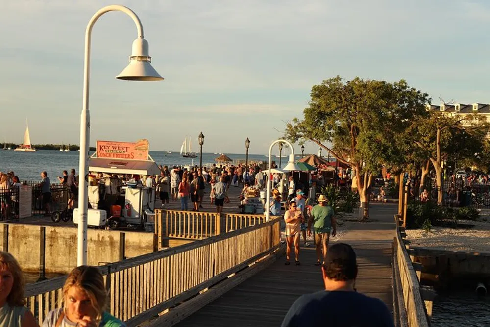 The image shows a bustling boardwalk by the sea with people walking and enjoying the evening and a sign that reads Key West Conch Fritters suggesting the scene is a lively area in Key West