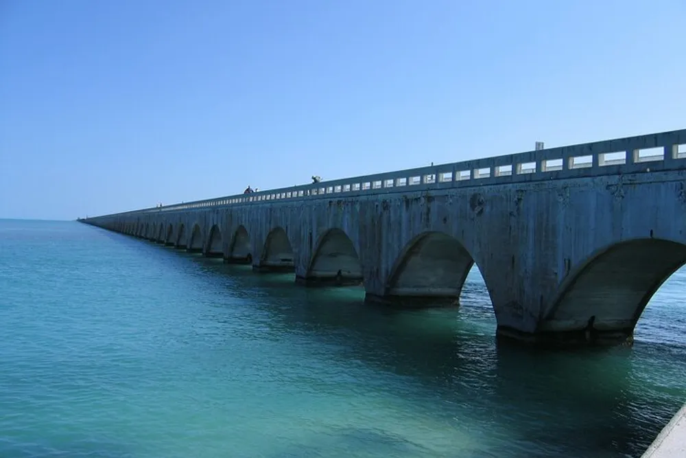 An arched concrete bridge extends over calm turquoise waters into the horizon under a clear blue sky