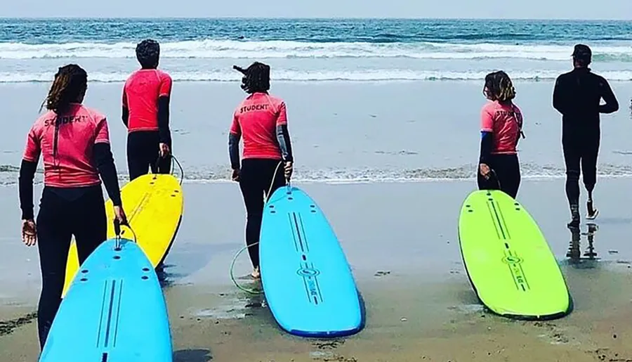 A group of surf students in colorful wetsuits and with surfboards are standing on the beach looking at the ocean.