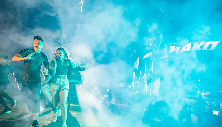 A group of people is running through fog with expressions of excitement, with bright lights and the word MAKO visible in the background.