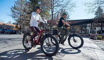 Two people are riding electric bicycles on a sunny day in an outdoor setting with shops in the background.