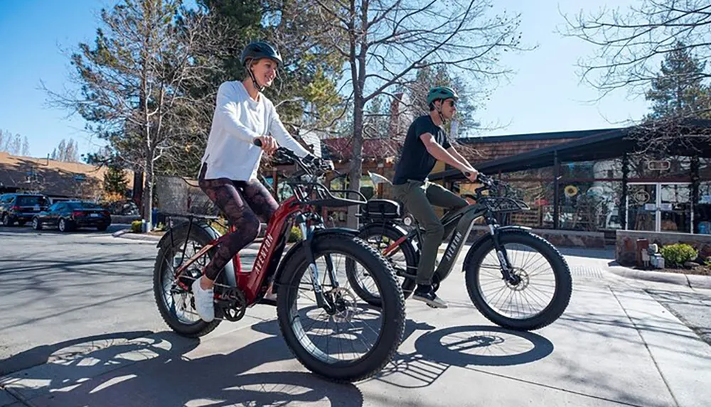 Two people are riding electric bicycles on a sunny day in an outdoor setting with shops in the background