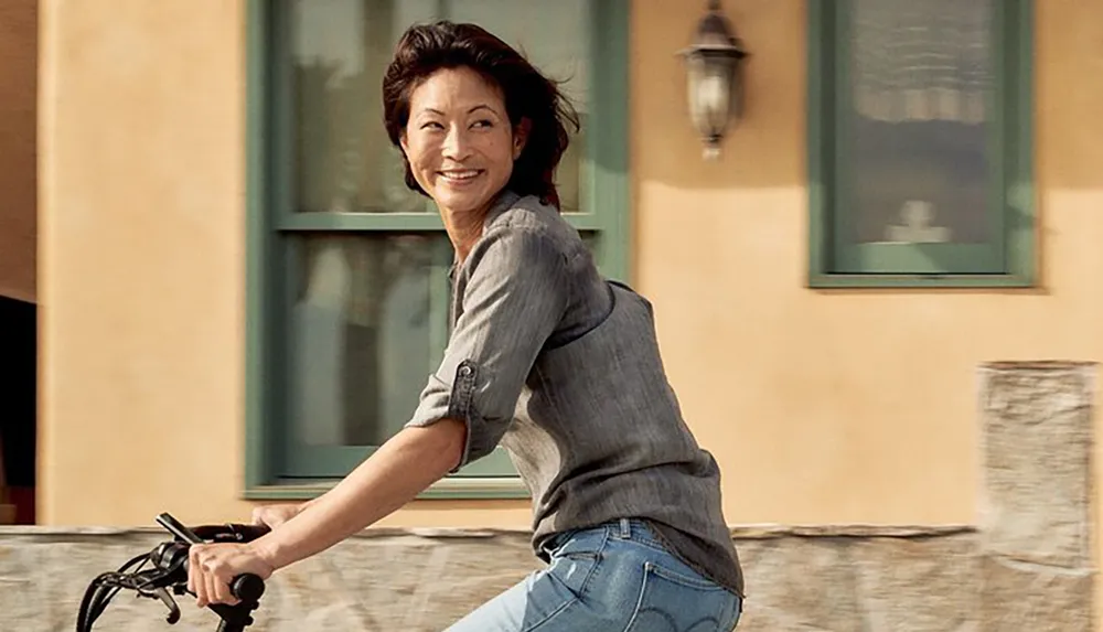 A smiling woman is enjoying a bicycle ride near a building with a vintage lantern and green windows