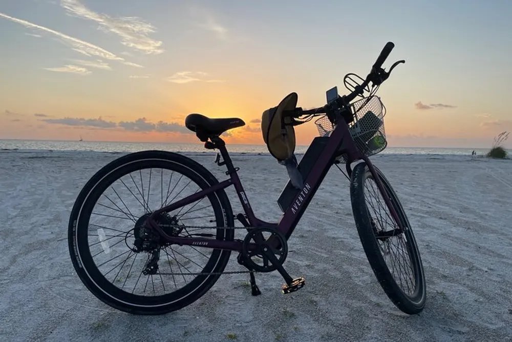 A bicycle with a hat on the handlebar is parked on a sandy beach against the backdrop of a sunset