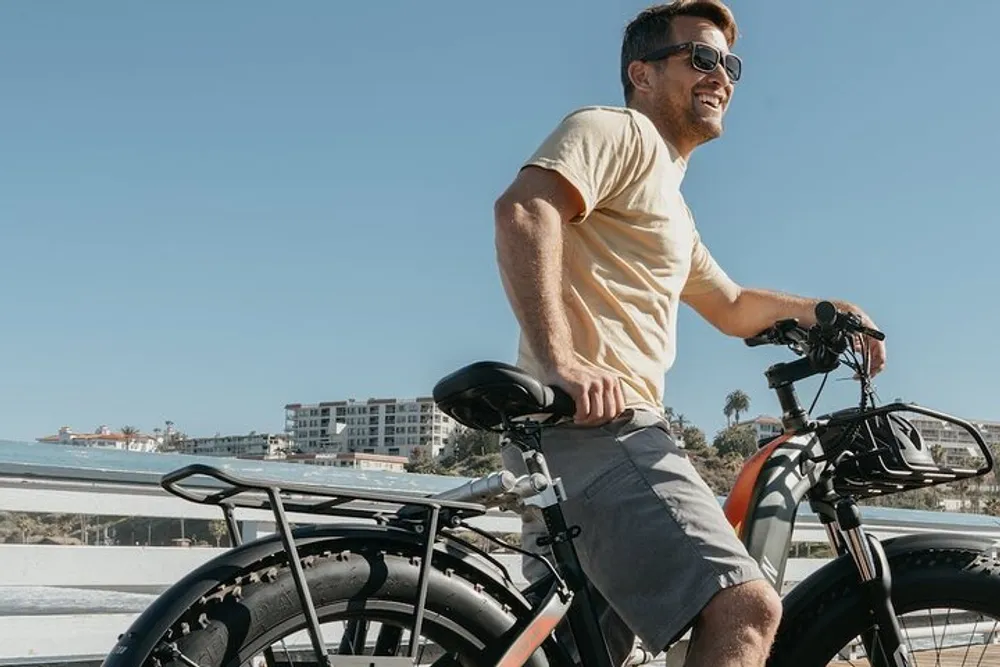 A smiling man is riding an electric bicycle along a sunny coastal pathway