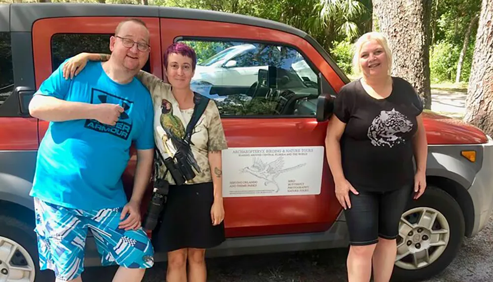 Three smiling individuals are posing for a photo next to a vehicle promoting birding and nature tours