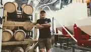 A man stands confidently with his arms crossed in a brewery or distillery, surrounded by wooden barrels and brewing equipment.