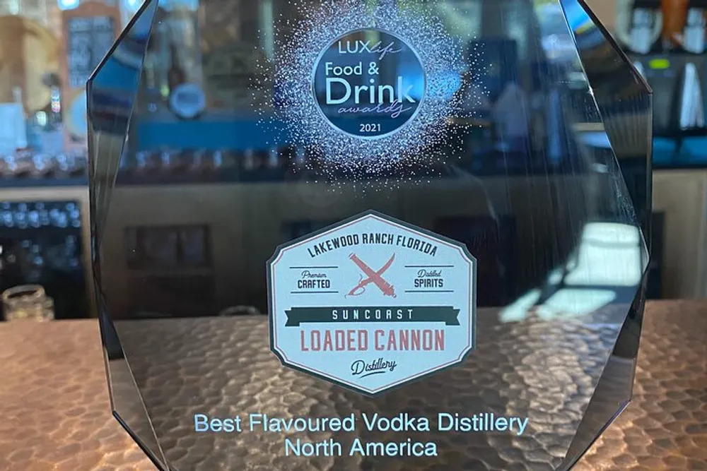 The image shows a clear glass award for Best Flavoured Vodka Distillery North America from LUX Life Food  Drink Awards 2021 sitting on a bar counter representing an accolade received by the Suncoast Loaded Cannon Distillery based in Lakewood Ranch Florida