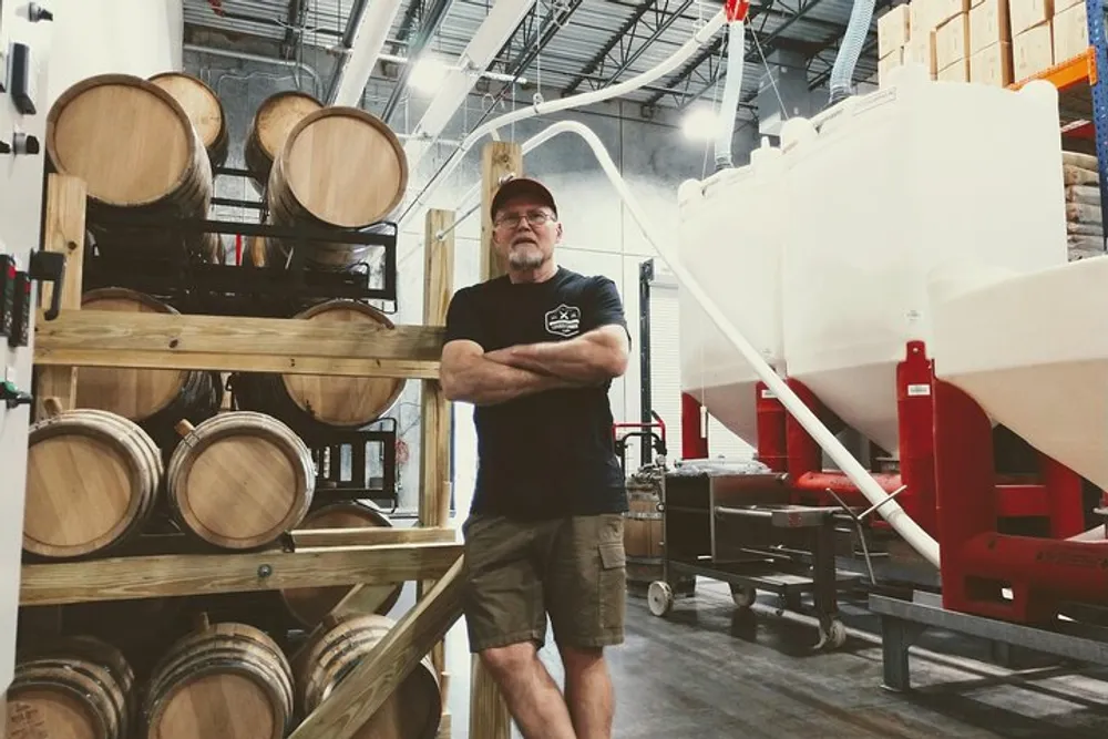 A man is standing with his arms crossed in a brewery or distillery environment surrounded by wooden barrels and brewing equipment