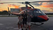 Three people are posing with thumbs up in front of an orange and black helicopter at sunset.