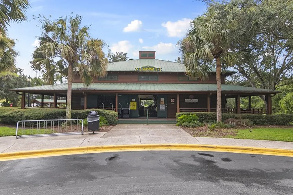 The image shows a single-story green building possibly a restaurant or visitor center with a covered porch area surrounded by trees and palms under a cloudy sky