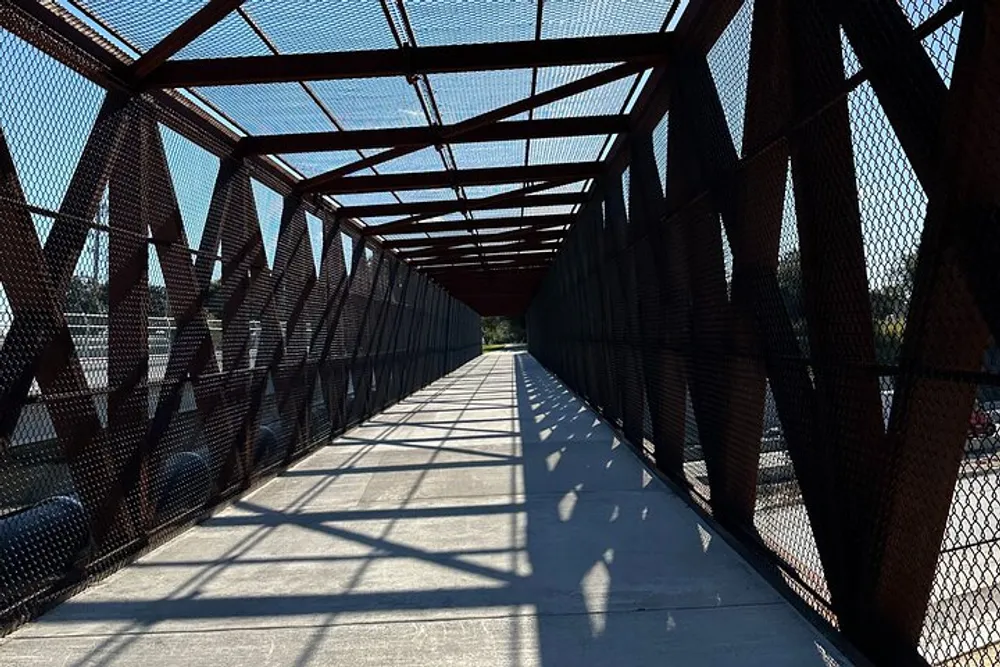 The image shows a long pedestrian walkway enclosed with a metal mesh and framed by a red truss structure with the sunlight casting geometric shadows on the ground