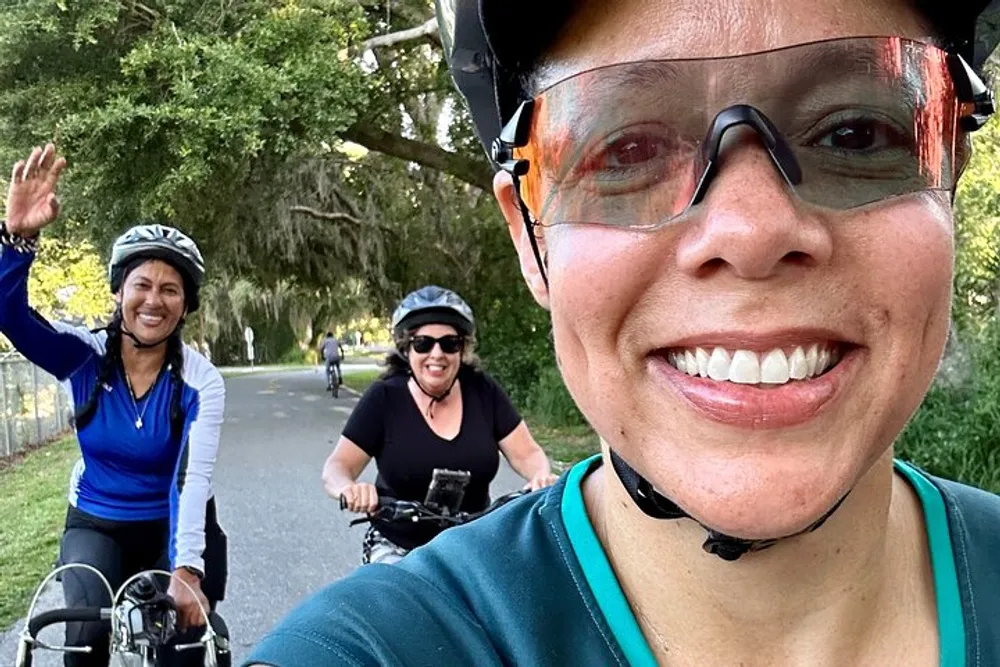A person in the foreground takes a selfie while cycling with two friends who are smiling and waving in the background on a tree-lined path