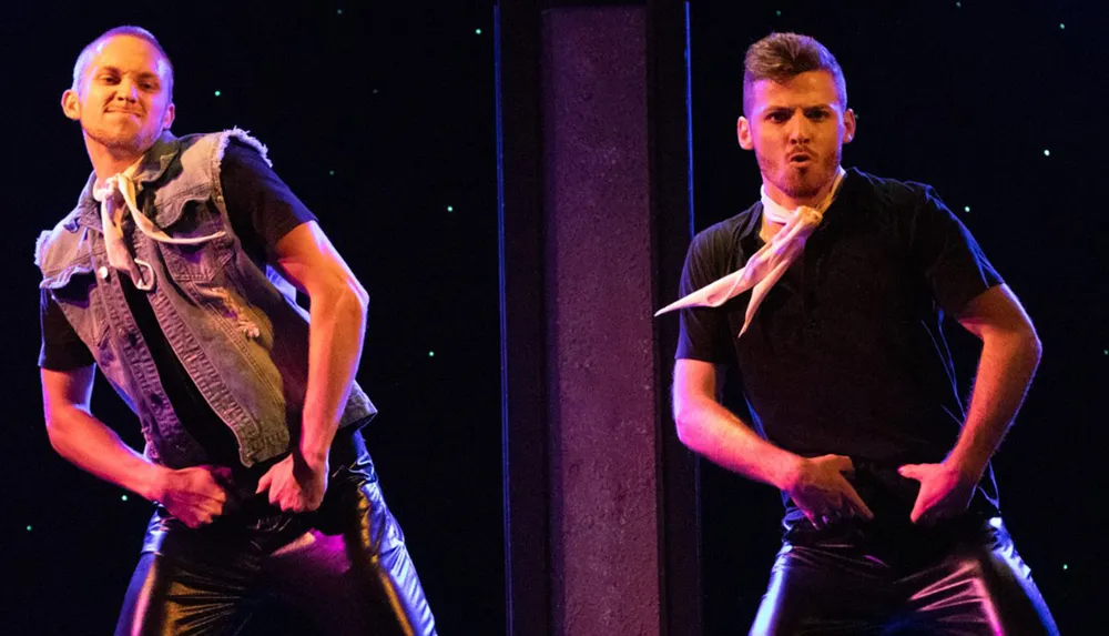 Two male dancers are performing with expressive facial expressions and dynamic poses on a stage with a dark backdrop speckled with small light points