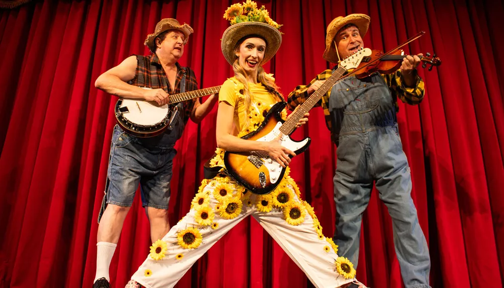Three performers dressed in exaggerated country-style clothing are playfully posing on stage with musical instruments against a red curtain backdrop