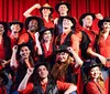 A group of performers dressed in red shirts black hats and country-western attire pose on stage with a red curtain backdrop smiling enthusiastically