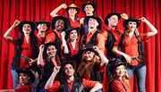 A group of performers dressed in red shirts, black hats, and country-western attire pose on stage with a red curtain backdrop, smiling enthusiastically.