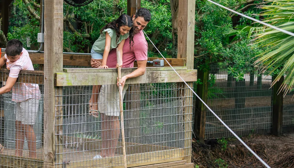 A man and a girl share a moment leaning over a wooden barrier at an outdoor enclosure while another child looks on from the side