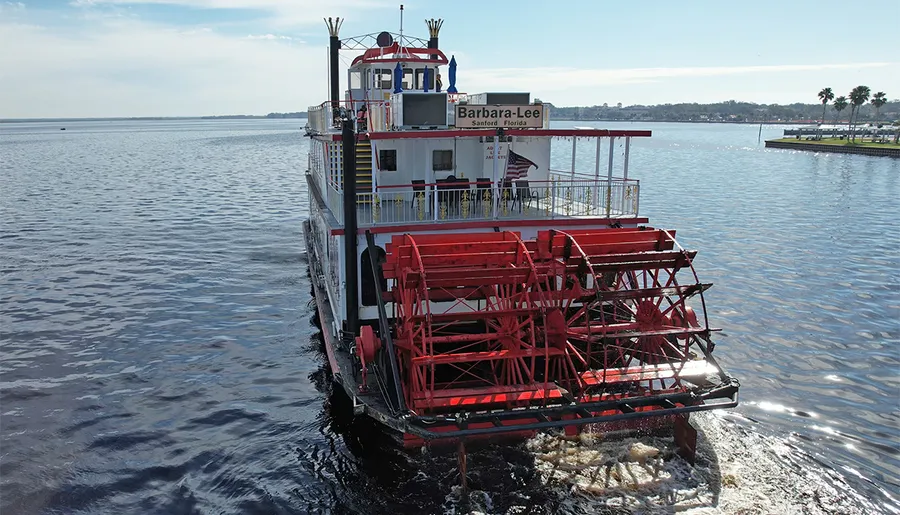 The image shows the stern of a red paddlewheel riverboat named 'Barbara-Lee' from Sanford, Florida, moving through the water.