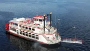 A traditional paddlewheel riverboat is cruising on calm waters under a clear sky.
