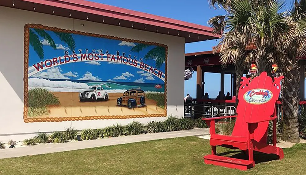 This image features an oversized mural proclaiming Daytona Beach as the Worlds Most Famous Beach with vintage cars on the shore adjacent to a large red Adirondack chair with branding outside a casual dining establishment